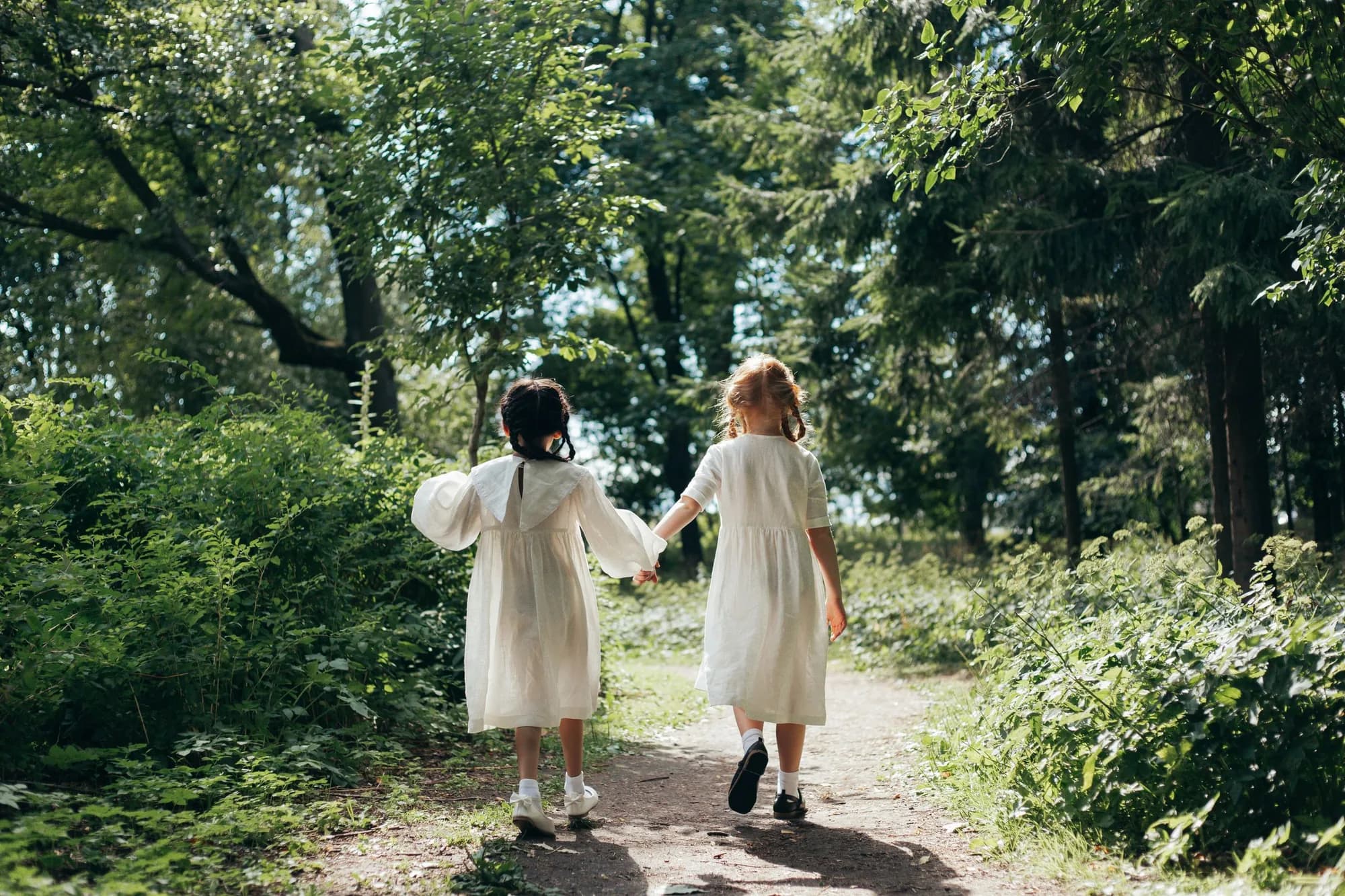 Two girls in white dresses walking hand in hand through a sunlit forest